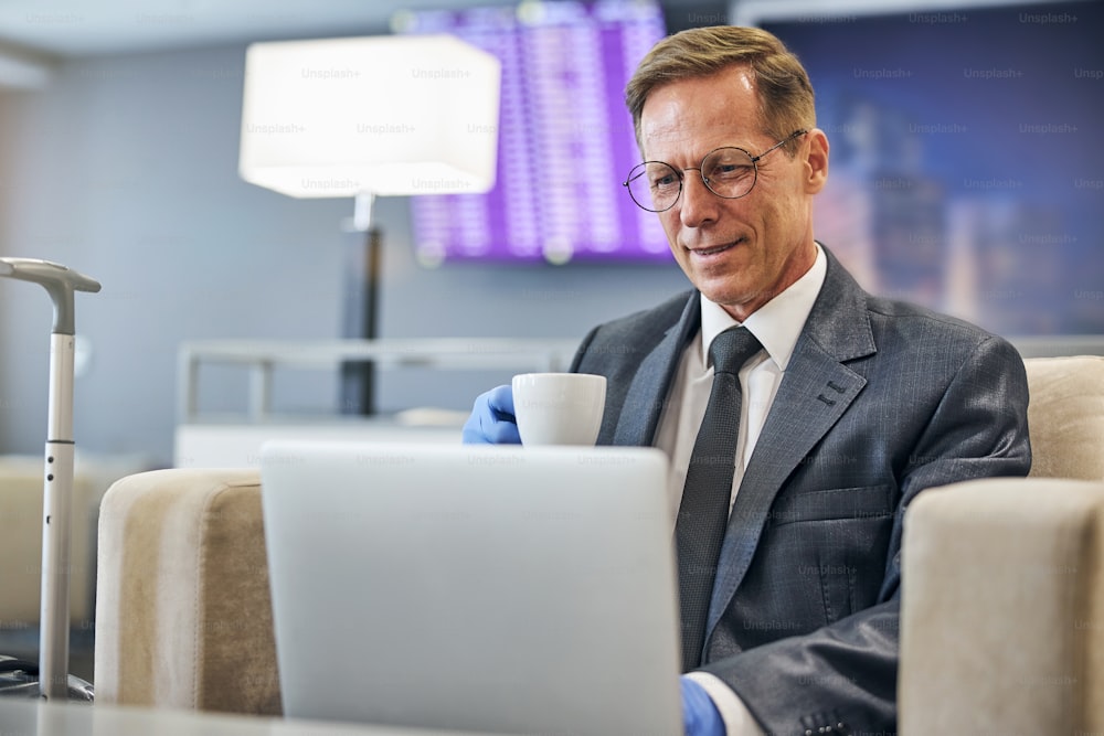 Cheerful elegant male in latex gloves and glasses is drinking coffee and using notebook before flight