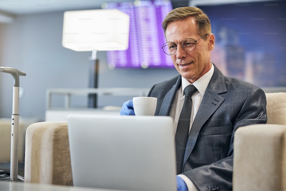 Fröhlicher eleganter Mann in Latexhandschuhen und Brille trinkt Kaffee und benutzt Notebook vor dem Flug