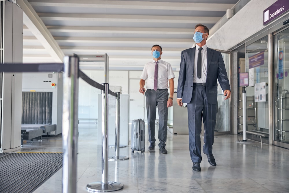 Elegant mature male is going on trip while his assistant is carrying luggage and they are wearing masks