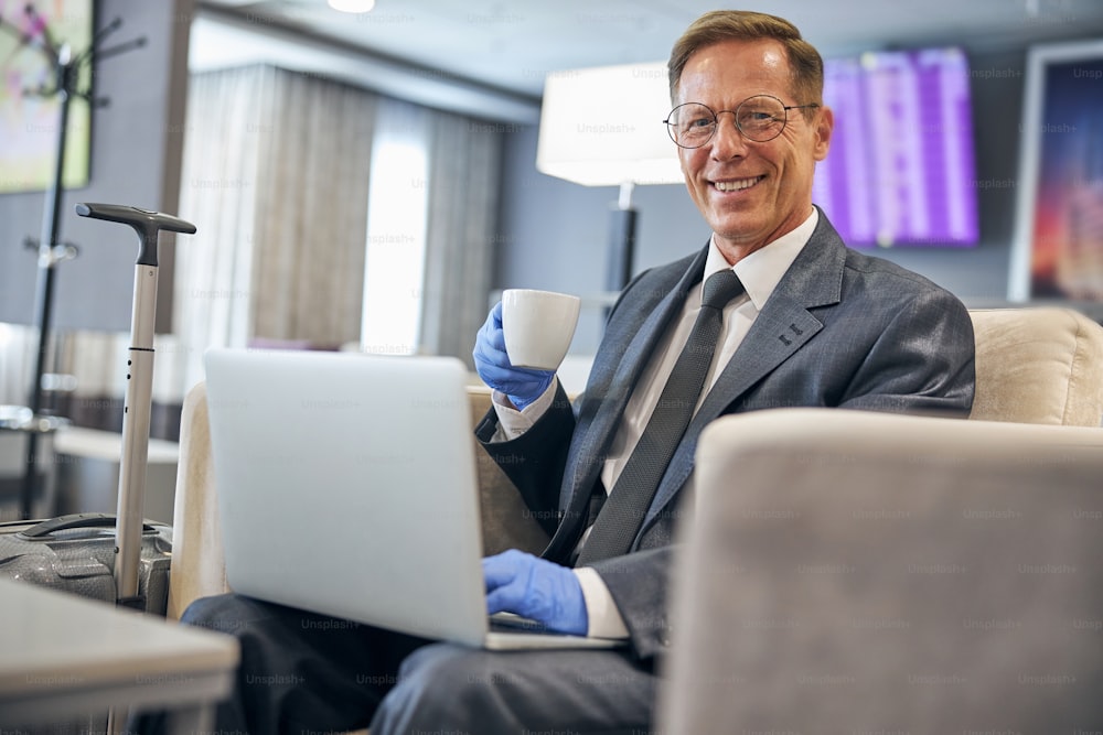 Ritratto dalla vita di un uomo d'affari elegante sorridente che usa il taccuino mentre beve caffè in guanti di lattice in aeroporto