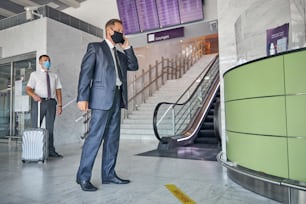 Elegante hombre maduro con máscara está haciendo una llamada mientras espera la salida con asistencia y llevando equipaje