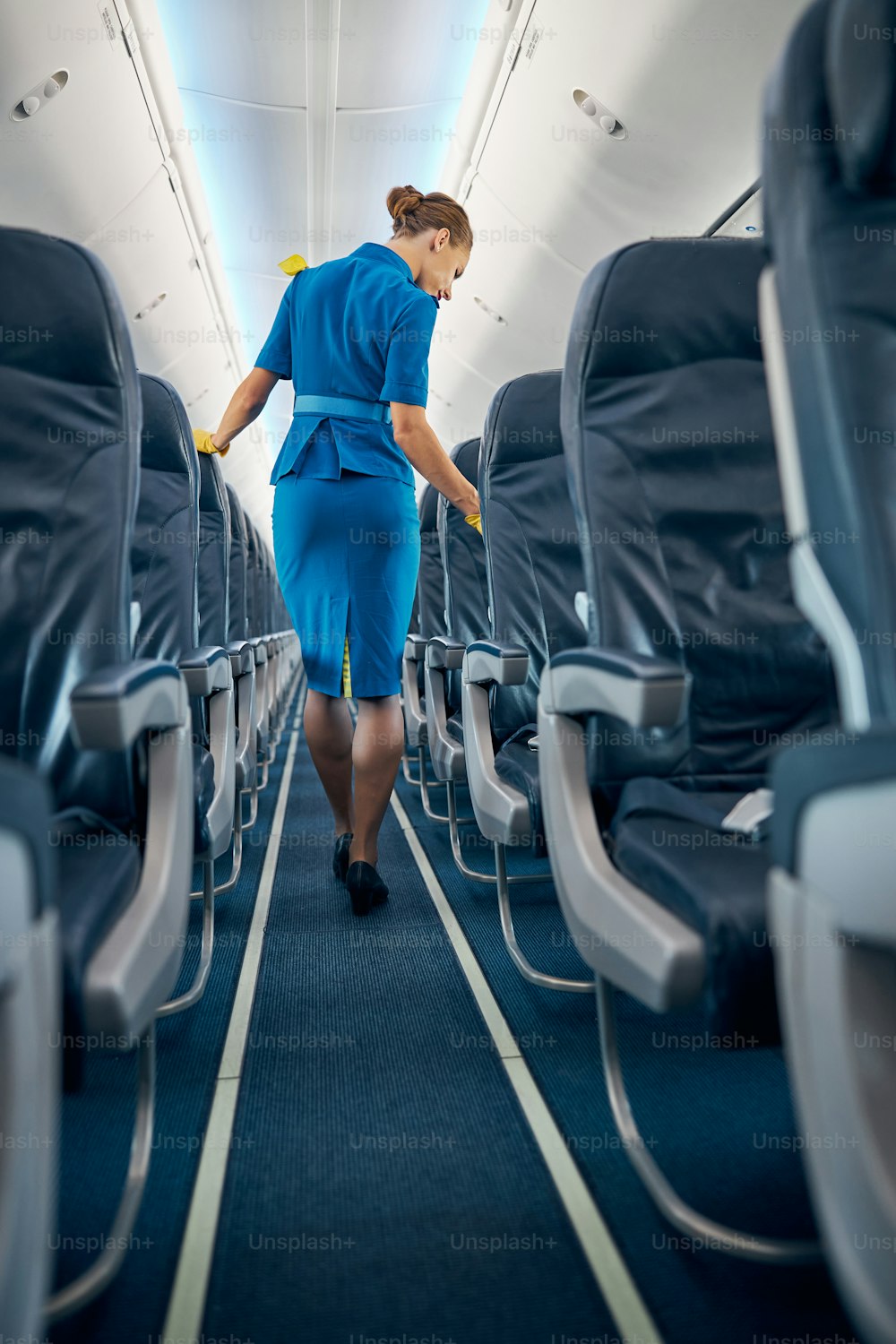 Back view portrait of joyful female in flight attendant blue uniform looking through the empty cozy passenger chairs