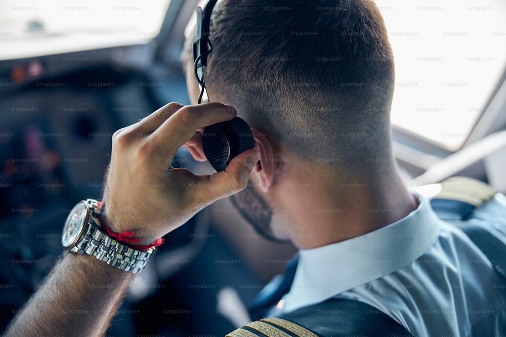 Back view portrait of bearded pilot with watch on hand while touching his headphone in the cockpit of aircraft