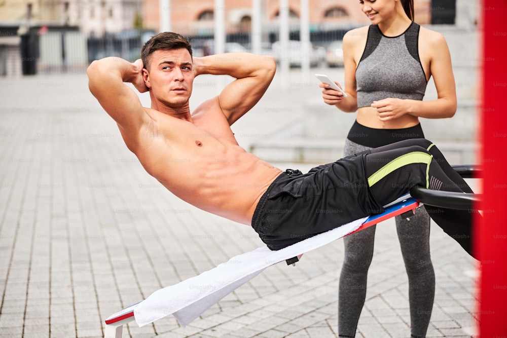 Shapely handsome male is doing crunches on bench on urban sports ground while slim woman is using smartphone