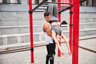 Un jeune homme musclé soutient une femme mince bien-aimée suspendue à une barre horizontale vers le haut sur un terrain de sport