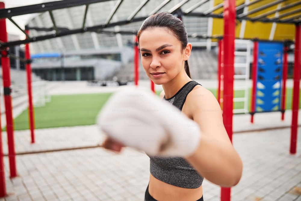 Waist up portrait of determined slim female having boxing training on sports ground outdoors and demonstrating punch
