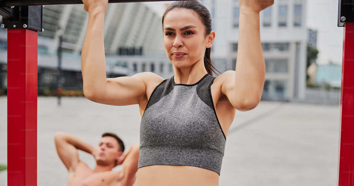 Pretty Abs Pretty slim female is doing pull ups on horizontal bar while muscular ...