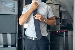 Close up of airline pilot in white shirt fixing his tie while standing inside the plane