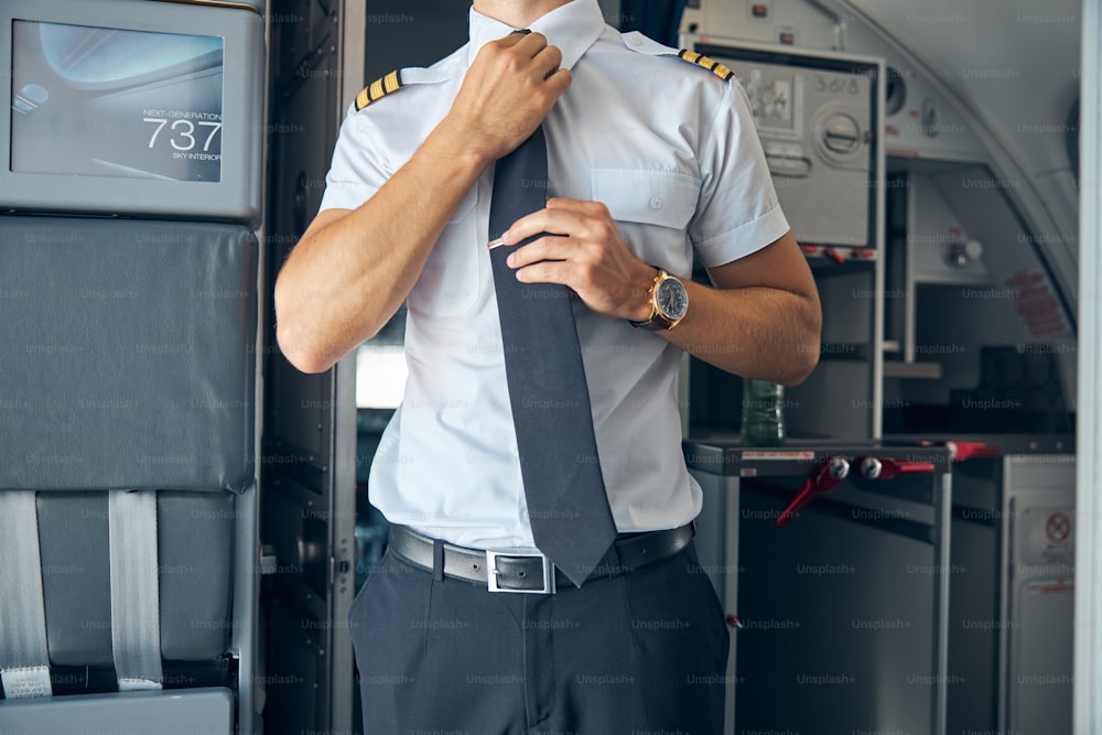 Close up of airline pilot in white shirt fixing his tie while standing inside the plane