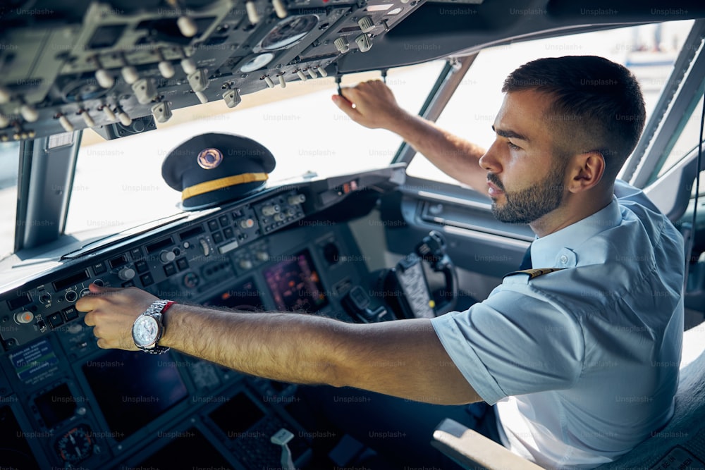 Retrato de la vista lateral de cerca guapo hombre confiado en uniforme sentado en la silla mientras él está comprobando el sistema del avión