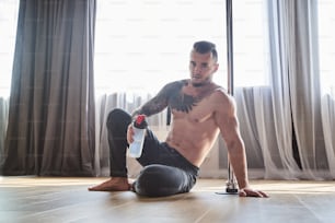 Retrato de cuerpo entero de un hombre musculoso guapo sentado en el piso de madera mientras sostiene una botella de agua en la mano en la clase de gimnasia