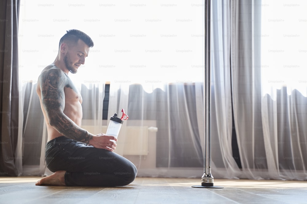Portrait of sporty male near pole dance sitting while holding bottle of water in hands