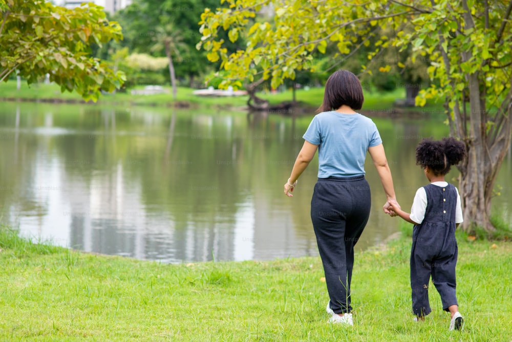 Vista posteriore della madre felice di razza mista con la figlia piccola che si tiene per mano e cammina insieme nel parco.
