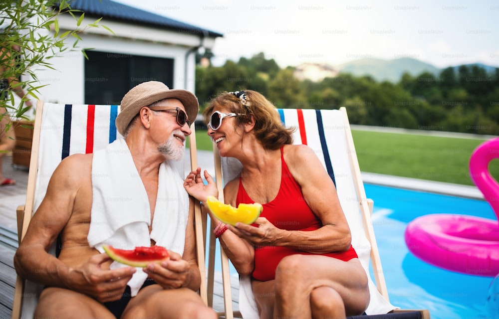 Cheerful senior couple sitting by swimming pool outdoors in backyard, talking.