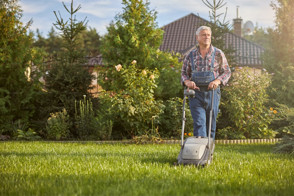 Ganzkörperfoto eines älteren Mannes, der mit einem Rasenmäher das Gras auf seinem Rasen trimmt