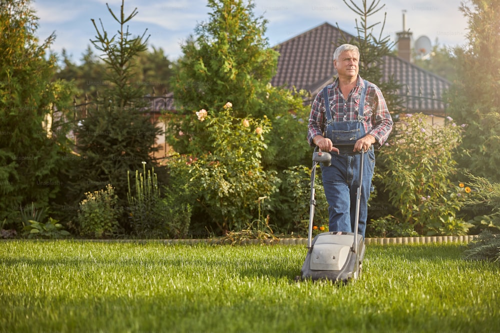 Photo en pied d’un homme âgé utilisant une tondeuse à gazon pour tondre l’herbe de sa pelouse