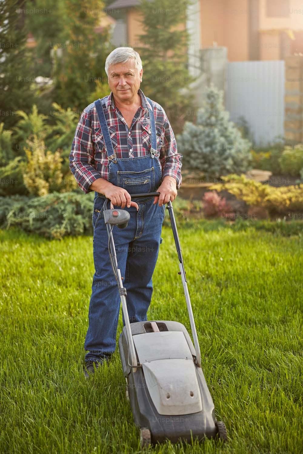 Full-length photo of a grey-haired man wearing a jeans jumpsuit and mowing his green lawn
