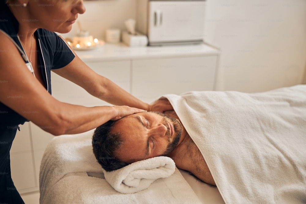 Handsome bearded gentleman with closed eyes receiving therapeutic massage while lying on massage table under white blanket