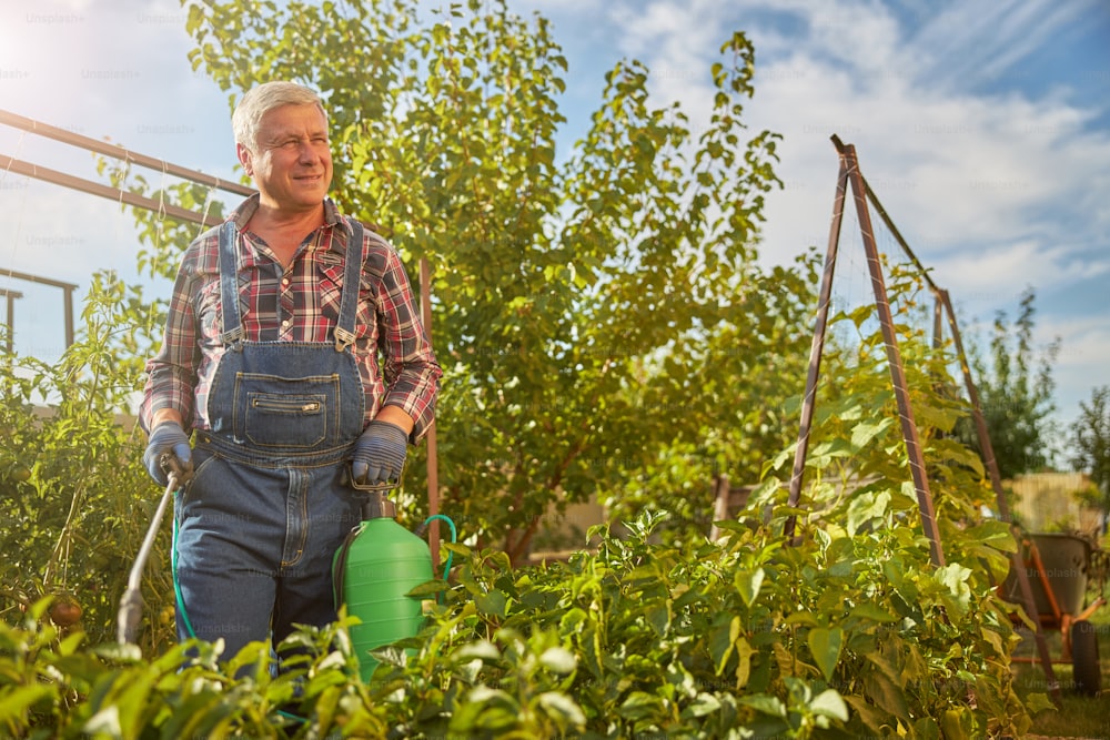 Cheerful aging man carrying a spray tank while sprinkling plants in his garden on a sunny day