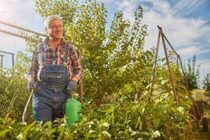 Hombre de edad alegre que lleva un tanque de pulverización mientras espolvorea plantas en su jardín en un día soleado