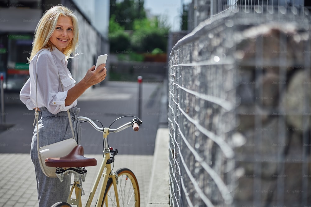 Waist up portrait of happy cheerful female in white blouse holding mobile phone while walking with bike on the city