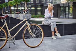 Full length portrait of stylish female crying in the outdoors while sitting behind of her bicycle in the modern city park