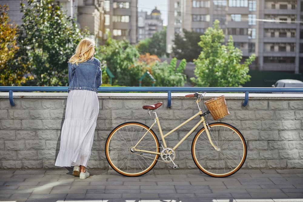 Mujer caucásica rubia adulta con vestido blanco y chaqueta de mezclilla de pie cerca de la bicicleta retro mientras toma un pequeño descanso de andar en bicicleta