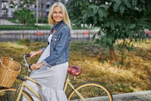 Side view portrait of attractive blonde Caucasian woman in stylish white long dress holding her hands on handlebar of city bike with basket
