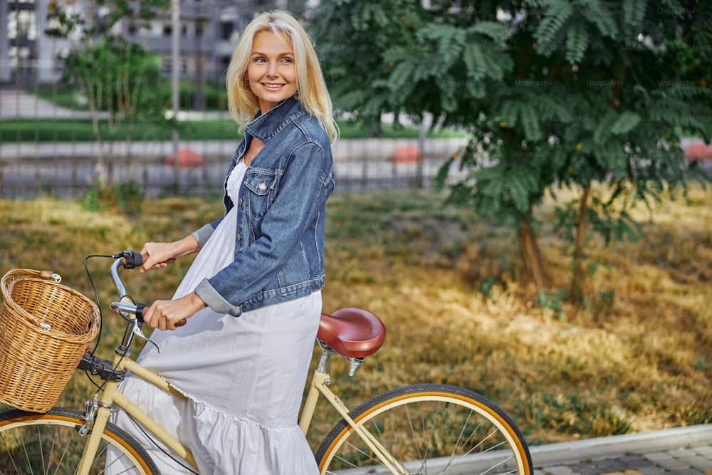 Side view portrait of attractive blonde Caucasian woman in stylish white long dress holding her hands on handlebar of city bike with basket