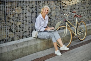 Full length portrait of happy smiling female in grey pants and white blouse working on laptop while sitting in the outdoors