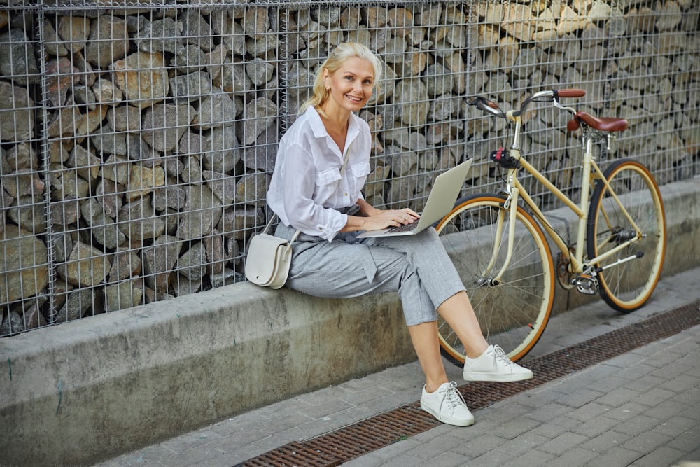 灰色のズボンと白いブラウスで幸せな笑顔の女性のフルレングスの肖像画は、屋外に座っている間、ラップトップで作業