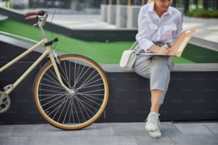 Cropped head portrait of stylish female in grey pants and white blouse writing something in the paper folder while bicycle standing near her