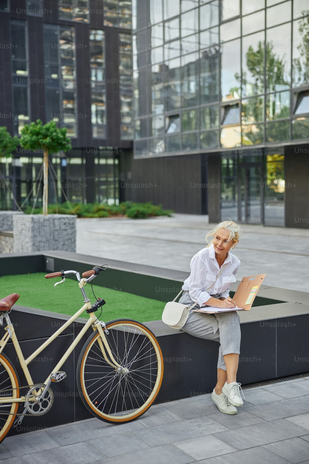 Full length portrait of good looking female in white clothes with modern bicycle sitting in front of the business center