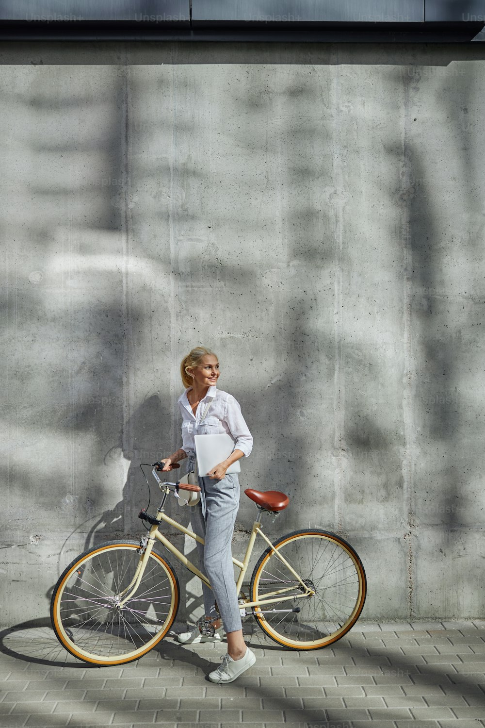 Portrait en pied d’une femme moderne avec un ordinateur portable gris à la main tout en passant du temps dans la ville vide avec son vélo rétro