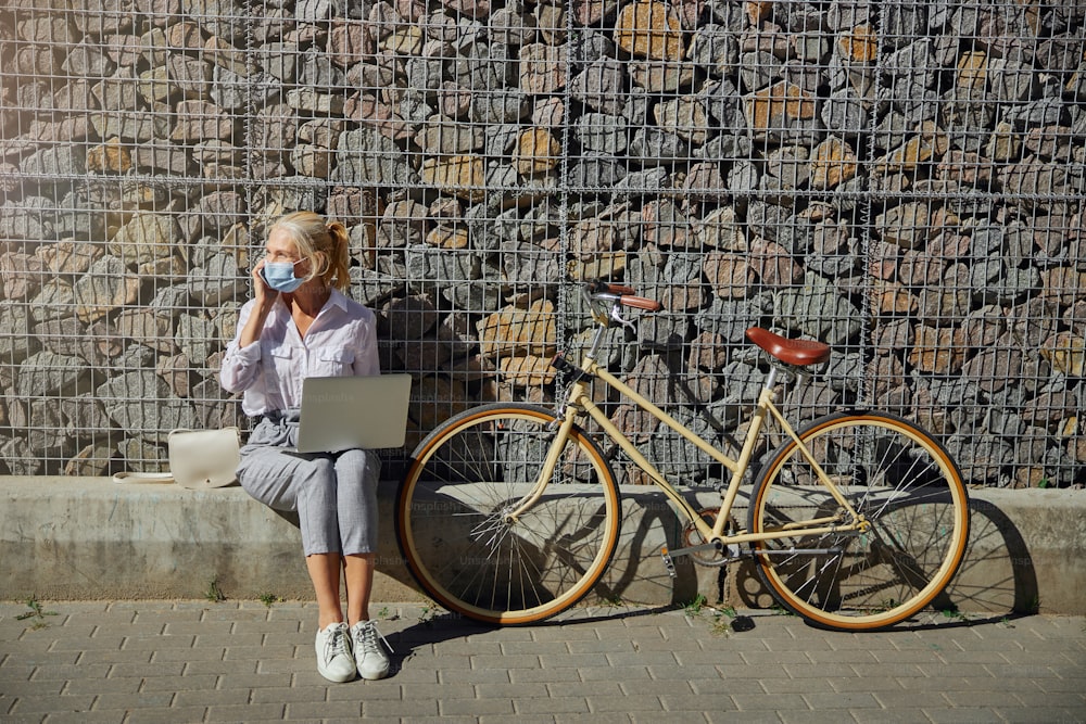 Mujer de moda con blusa blanca y pantalones grises con bicicleta retro que pasa tiempo en la ciudad aislada en la pared de piedra