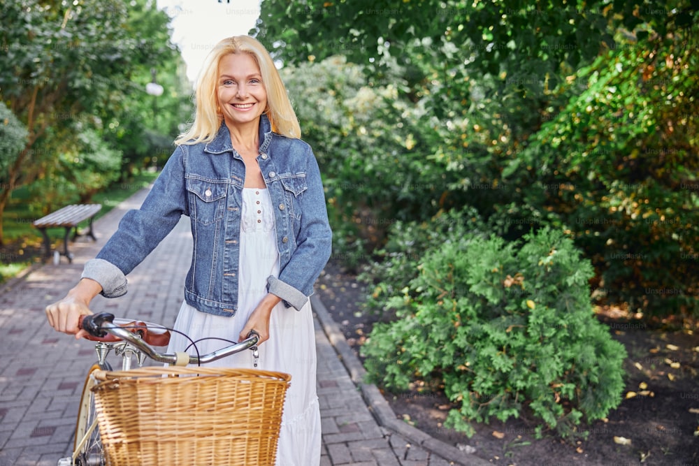 Close up portrait of happy cheerful lady in denim jacket and white dress holding a handlebar of a city bike with basket