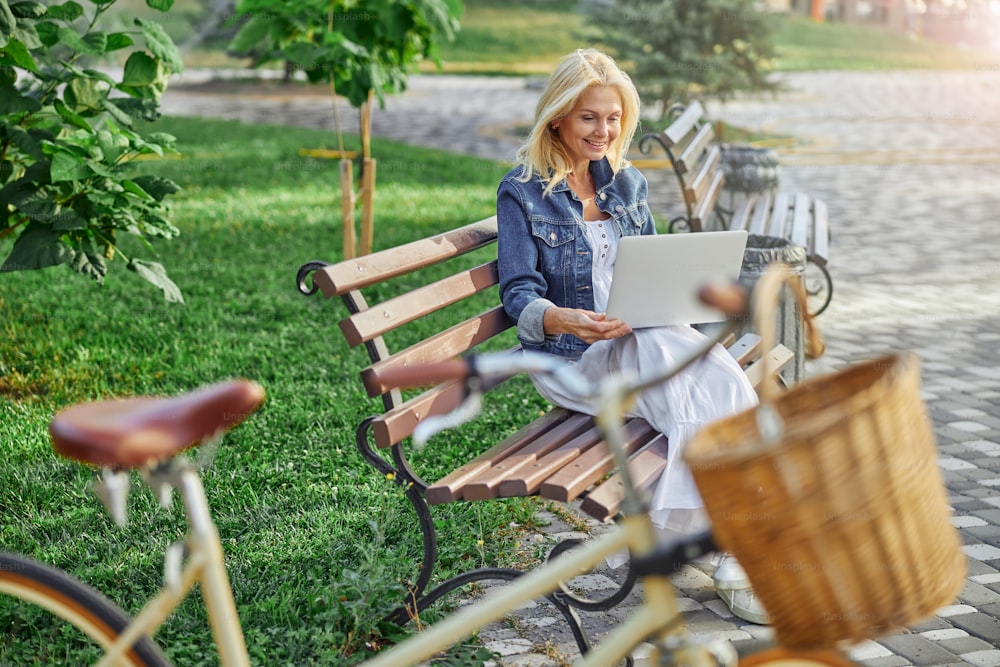 Retrato da vista lateral da mulher adulta sorridente feliz sentada no banco enquanto trabalha no laptop moderno no parque da cidade