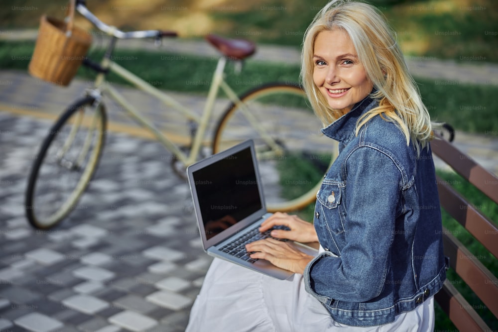 Close up portrait of happy cheerful woman in denim jacket looking at the photo camera while sitting on the wooden bench in front of bicycle