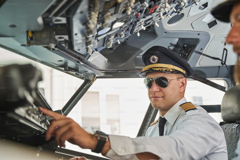 Attractive pilot in white uniform and black sunglasses putting one hand forward and pushing a button on a control panel in cockpit