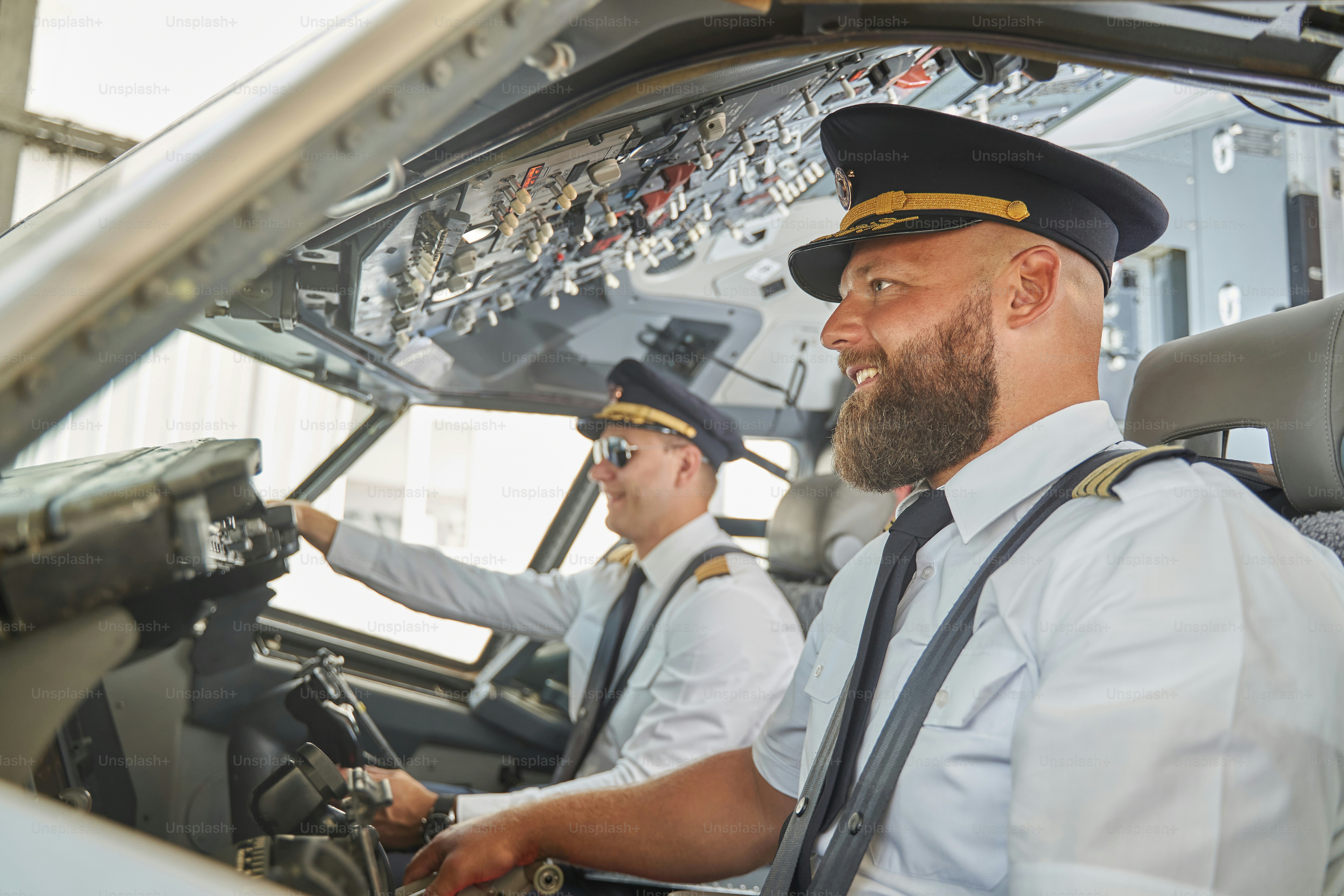 Due piloti attraenti positivi che sorridono nella cabina di pilotaggio dell'aereo mentre sono seduti con le cinture di sicurezza allacciate
