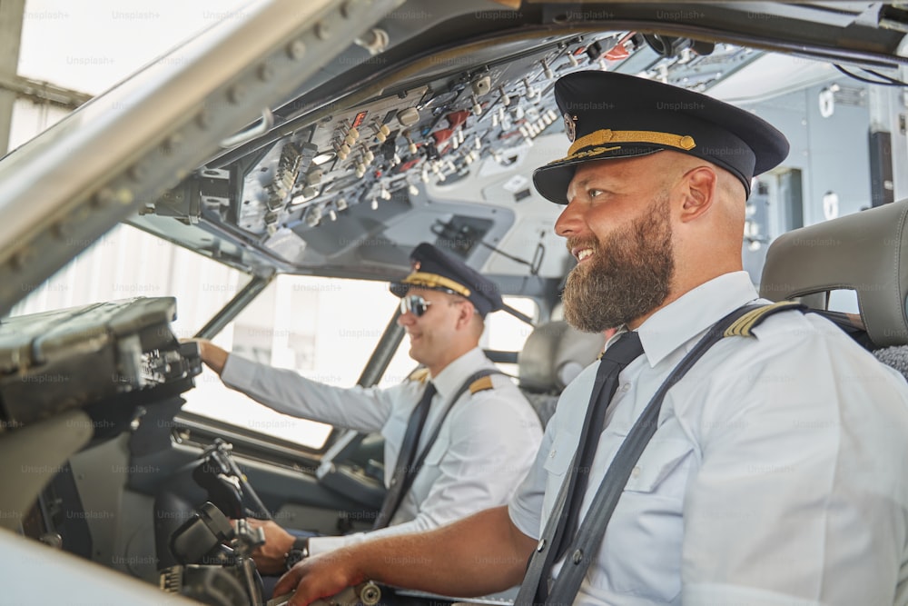 Dois pilotos positivos e atraentes sorrindo no cockpit do avião enquanto estão sentados com os cintos de segurança apertados