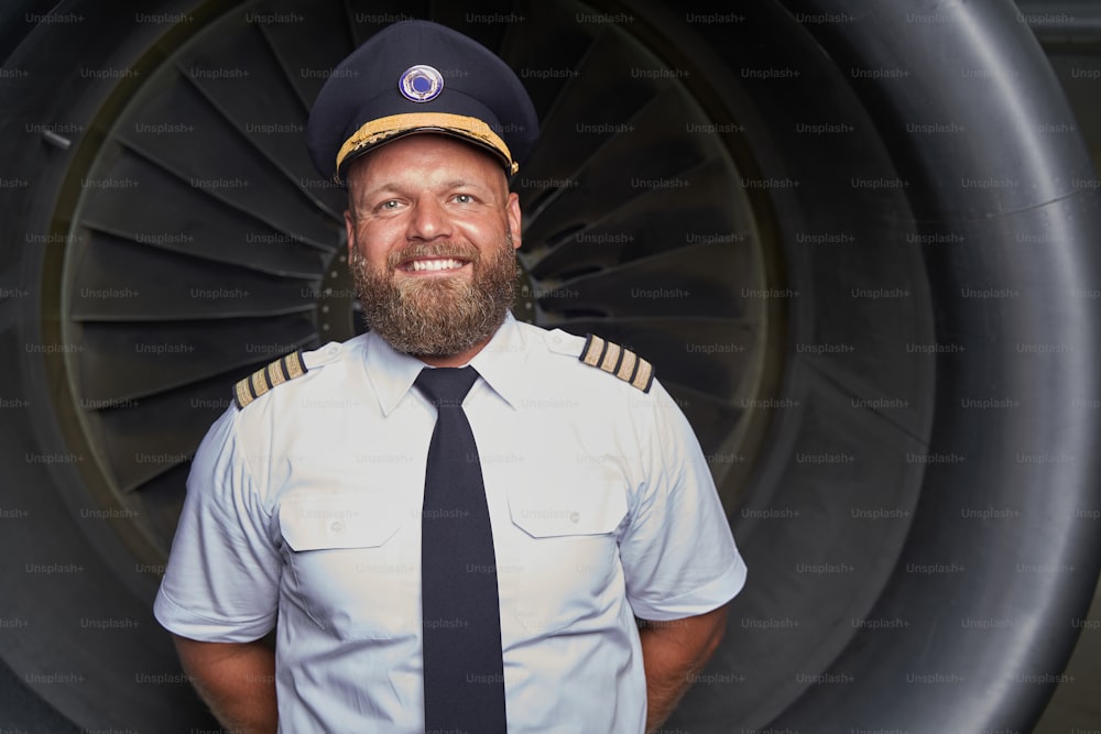 Joyeux aviateur barbu en uniforme blanc debout et souriant. Un moteur à réaction dans le dos