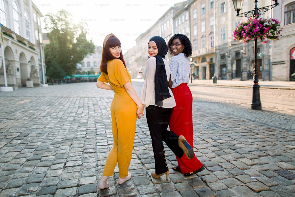 Back view of three diverse young female laughing friends, wearing stylish colorful clothes, walking arm in arm together in the city at sunny day. Pretty Caucasian, African and Muslim girls in city