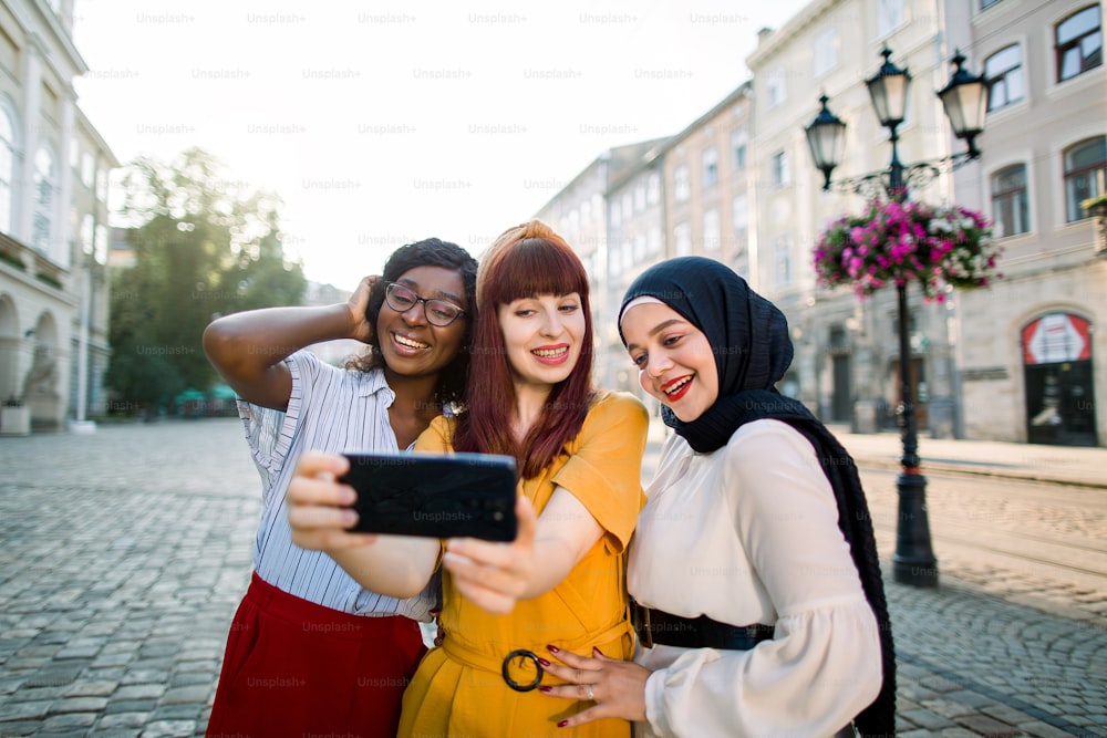 Grupo multirracial de tres jóvenes atractivas mejores amigas con ropa colorida, tomando selfie en una hermosa calle urbana. Tres mujeres jóvenes, africanas, musulmanas y caucásicas caminando al aire libre en la ciudad.