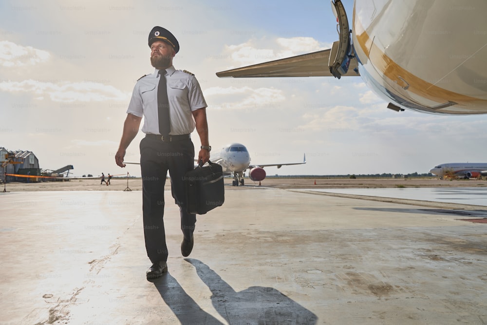 Confident bearded pilot carrying a briefcase and looking into the distance while walking toward the airplane