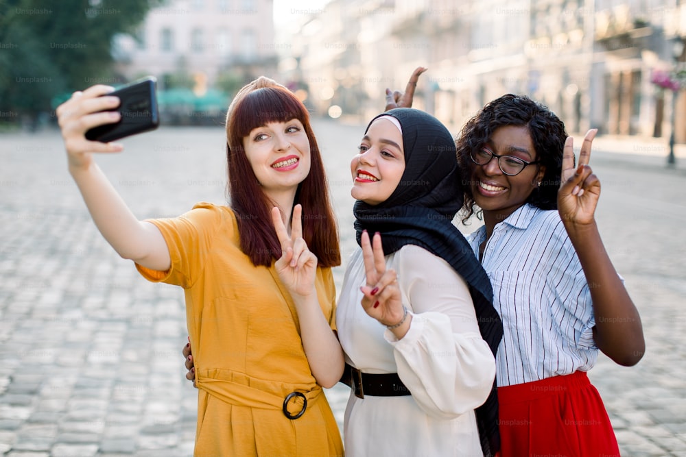 Mejores amigas, tres chicas multirraciales con teléfono inteligente, riendo, gesticulando el signo de la paz y haciéndose una foto selfie. Tres mujeres en una ciudad europea caminando por la calle. Tecnología y conceptos de amistad.