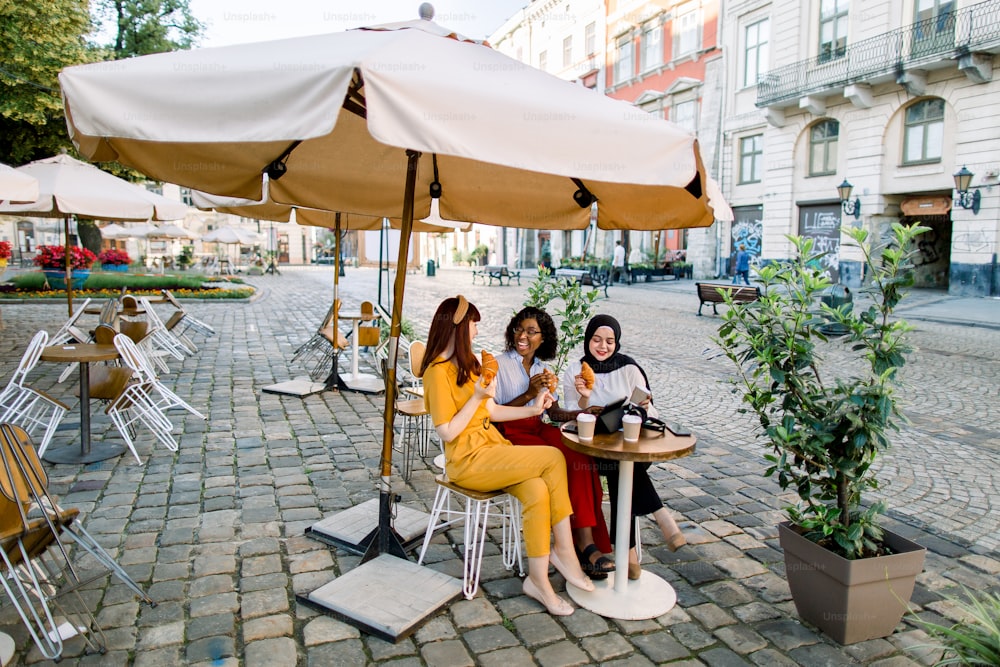 Urban lifestyle, lunch time, friendship concept. Three attractive smiling young diverse women friends talking and laughing, drinking coffee with croissants while sitting at table in cafe outdoor.