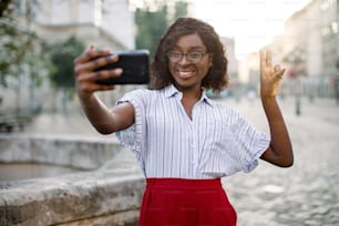 Colpo urbano all'aperto di bella giovane donna d'affari sorridente afroamericana, che fa segno di pace e autoritratto su smartphone, in posa nella vecchia strada della città. Selfie, persone, concetto di stile di vita