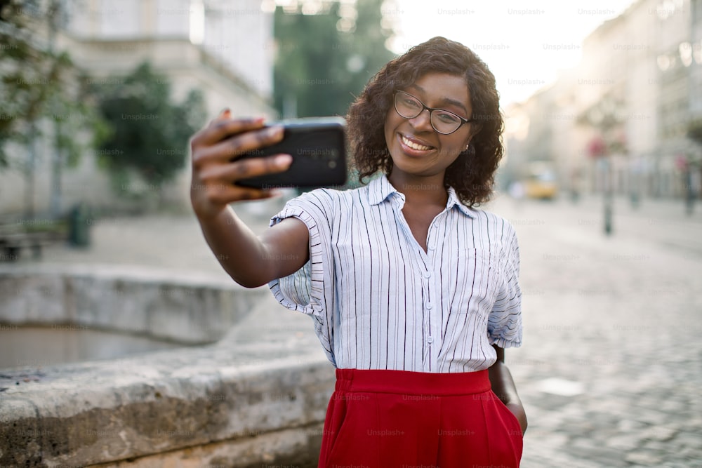 Portrait of cheerful young african american business woman,wearing red pants and striped shirt, taking selfie on her smartphone outdoors, posing in old city street near ancient stone fountain.