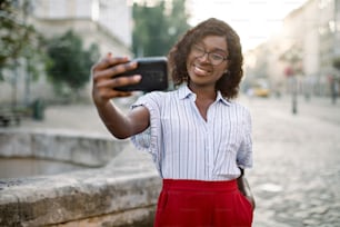 Ritratto di allegra giovane donna d'affari afroamericana, che indossa pantaloni rossi e camicia a righe, che scatta selfie sul suo smartphone all'aperto, in posa nella vecchia strada della città vicino a un'antica fontana di pietra.